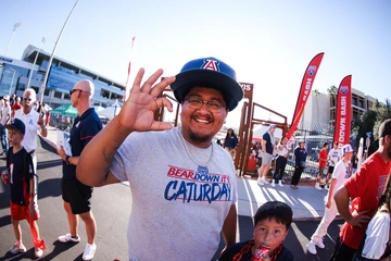 A smiling University of Arizona fan wearing a “Bear Down It’s Caturday” T-shirt and oversized Wildcat hat flashes the “OK” hand sign while standing outside Arizona Stadium during a Bear Down Bash event. Other attendees, including a child with a soda, are visible in the background amid red banners and festive crowds.