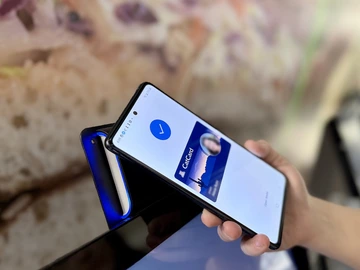 A person holds a smartphone displaying a digital CatCard in a mobile wallet app, tapping it against a card reader with a blue light to gain access.