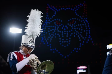 A University of Arizona marching band member in a red, white and blue uniform plays a brass instrument under bright stadium lights at night. Behind them, hundreds of red and blue drones form the image of the university’s Wildcat logo in the sky.