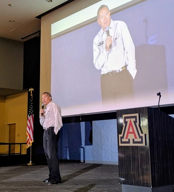 Interim Chief Information Officer Elliott Cheu speaks on stage at an IT summit, holding a microphone beside a podium with the University of Arizona logo. A large projection screen behind him displays his image as he addresses the audience.