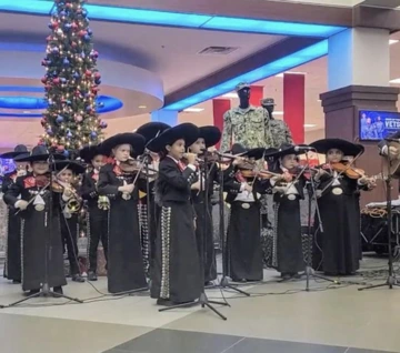 A youth mariachi group performs live at the University of Arizona’s Festivus event, standing in front of microphones and playing violins and trumpets in traditional black mariachi attire with red ties. A decorated Christmas tree glows behind them inside the Campus Store.