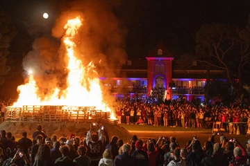A large bonfire blazes in front of Old Main during the University of Arizona Homecoming celebration. A crowd of people watches and takes photos as flames rise high into the night sky under a full moon.