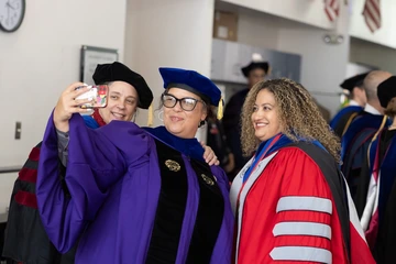 Three University of Arizona faculty members in academic regalia smile as they take a selfie together before a ceremony. One wears a purple gown, another a blue cap and gown, and the third a red gown. Other faculty in similar regalia are visible in the background.