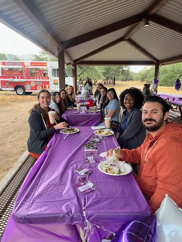 A group of people sat at a long picnic table under a metal-roofed pavilion. The table is covered with a purple plastic tablecloth, and there are plates of food and drinks in front of each person. The attendees appear to be enjoying a meal together. Purple balloons are tied to the table. Surrounding the pavilion is a grassy area with trees.