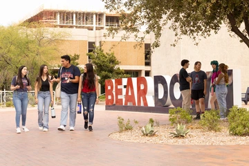 Students walk and talk near the large red, white and blue “BEAR DOWN” letters in the University of Arizona’s Student Success District. In the background, other students stand and chat under a tree, with campus buildings and a construction site visible behind them.