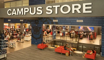 Interior view of the University of Arizona Campus Store showing displays of Wildcat apparel and merchandise, with shoppers browsing and people seated on red couches in the foreground.