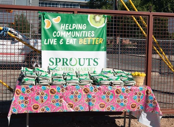 A colorful table covered with a bright floral cloth displays neatly stacked green-and-white tote bags in front of a banner reading “Helping communities live & eat better – Sprouts Farmers Market.” A small basket of lemons sits at one end of the table.