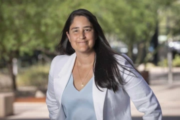 A woman in a lab coat stands near green trees.