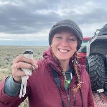 A woman wearing a red jacket and black beanie holds a small white bird in her right hand while smiling. She stands among a field of small brush, with the back of a truck barely visible behind her shoulder.