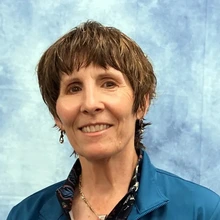 A woman with short brown hair, wearing a light blue button down, sits in front of a mottled blue and white background in a photography studio.