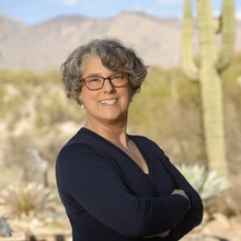 A woman wearing a black long sleeve shirt, with short curly grey hair, stands with her arms crossed in front of a field of desert plants and saguaro cacti.