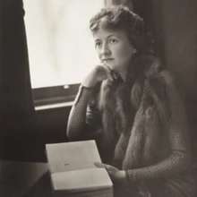 A black-and-white photograph of a woman sitting by a window. She has short, wavy hair and is wearing a patterned long-sleeve top with a fur stole draped over their shoulders. She is gazing out the window thoughtfully, with one hand resting on her chin.