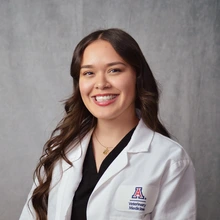A person with long, wavy brown hair, smiling broadly at the camera. They are wearing a white coat over a black top, and a necklace with a small pendant. The background is a textured gray, providing a neutral backdrop. The coat has a badge on the left side with a logo featuring an "A" and the text "Veterinary Medicine."