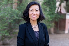 A woman with shoulder-length black hair smiles confidently in an outdoor setting with green trees in the background. She wears a black blazer over a patterned blouse and a silver necklace.