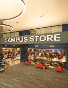 The entrance to the University of Arizona Campus Store inside the Student Union Memorial Center, with a large “Campus Store” sign above and racks of red-and-blue merchandise visible inside. In the foreground, several people sit on red couches in a lounge area.