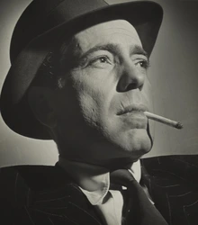 A dramatic black-and-white portrait of actor Humphrey Bogart wearing a fedora and pinstripe suit. He gazes upward and off to the side, with a cigarette resting between his lips. Strong lighting emphasizes the texture of his skin and the contours of his face.