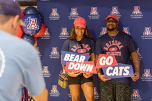Two people wearing Arizona Wildcats shirts and red hats pose in front of a University of Arizona backdrop at Destination Arizona, holding signs that read “Bear Down” and “Go Cats.”