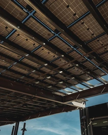 View looking up at a canopy of solar panels mounted on a metal framework, with sunlight shining through the grid-like pattern against a blue sky.