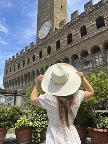 A student wearing a wide-brimmed hat and white dress looks up at a historic stone building with a large clock tower in Florence, Italy.