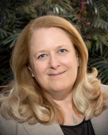 A woman with blonde hair and a tan blazer posing in front of trees.