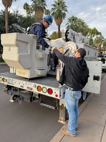 A Facilities Management employee wearing a hard hat climbs into the bucket of a utility truck while another worker on the ground hands him equipment. Palm trees and campus buildings line the street behind them.