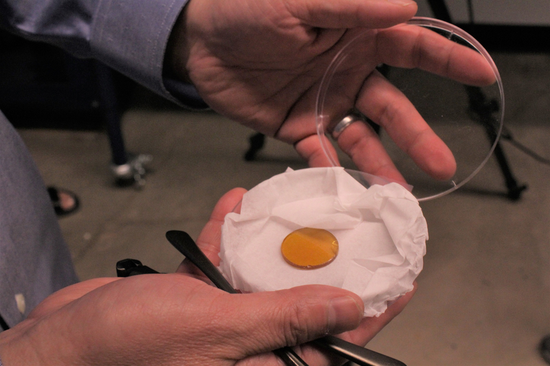 The orange material sits in a petri dish covered in a white cloth. A researcher pulls off the petri dish's lid to show the polymer