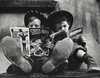Two boys in cowboy hats sit side by side with their feet stretched out toward the camera, each reading a comic book. One holds The Ghost Rider while the other reads a Zane Grey western. The image is in black and white, highlighting their worn shoes and absorbed expressions.