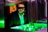 A man wearing safety glasses and a suit observes a green-lit optical experiment with lasers and mirrors in a laboratory setting.