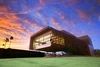 A modern, angular building with a geometric design situated against a dramatic sky during sunset. The structure features large glass windows framed by brown, asymmetric panels. The upper section of the building extends outward, creating an overhang supported by columns. The sky behind the building is a vibrant mix of orange and purple hues, with scattered clouds reflecting the sunset colors.