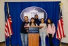 Five young people standing together behind a podium, which indicates "The White House Washington." The group is posed in front of a royal blue curtain displaying a large emblem of the White House. Two American flags are positioned on either side of the curtain, each with a gold-colored eagle finial at the top. The individuals are smiling and casually dressed, with one wearing a hijab.
