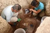 archaeologists crouching in a hole in the ground and digging around artifacts buried in the ground