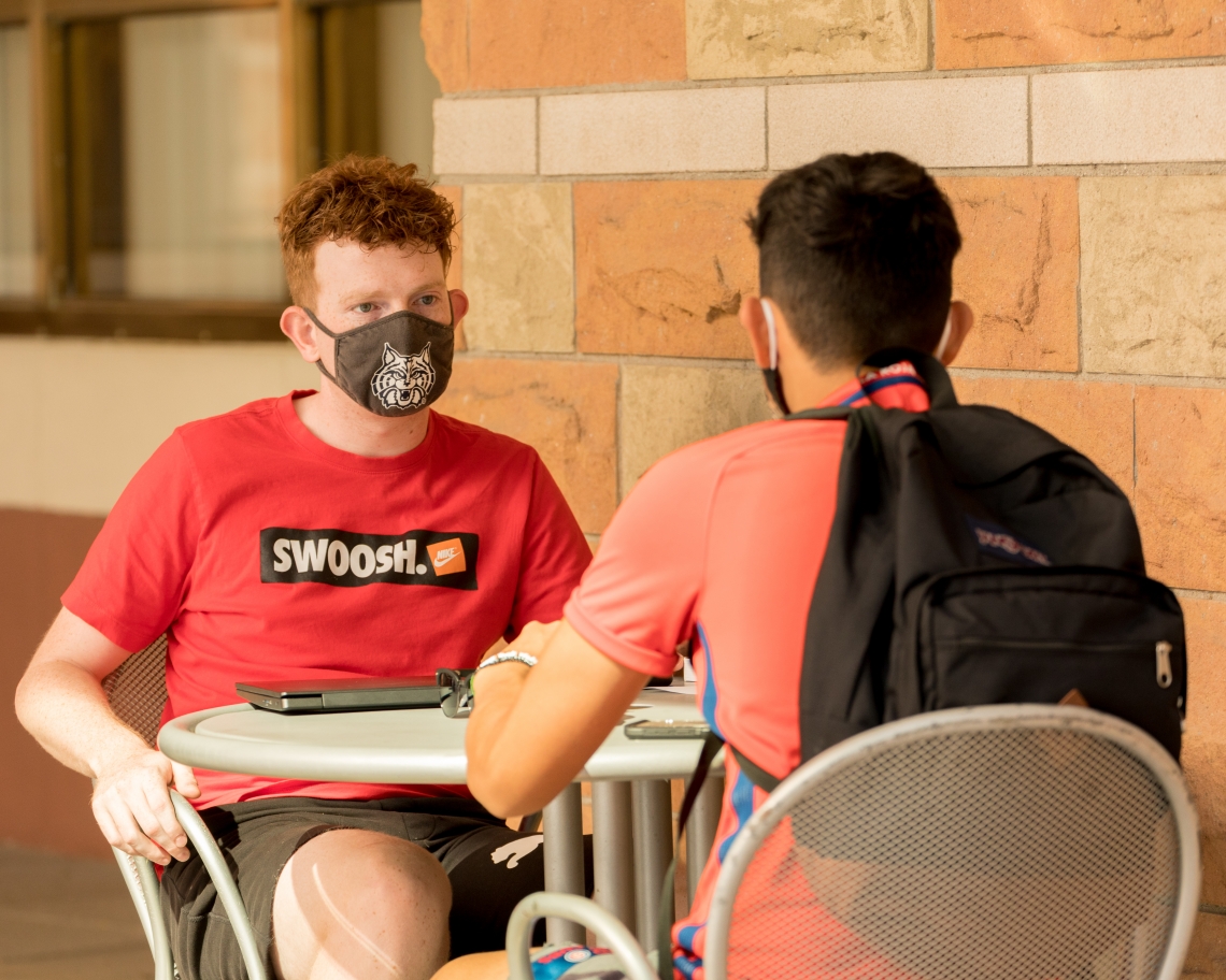 two male students sitting at an outdoor table wearing masks