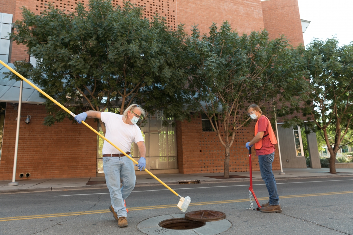 two men stand by a manhole, with one scooping a bottle of liquid from it