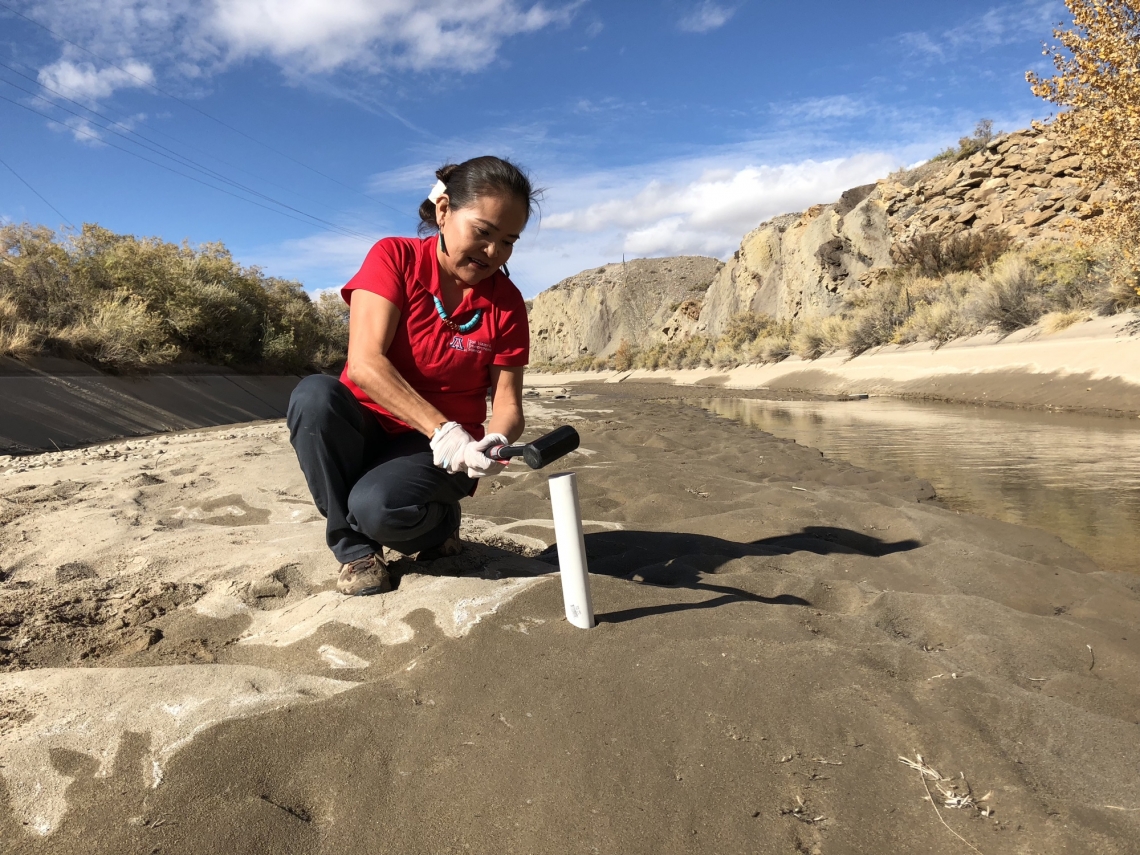 Native American woman uses a hammer to pound a pipe into the ground