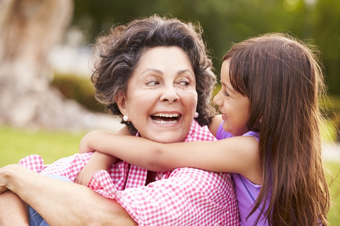 a seated older Hispanic woman smiles as a young girl embraces her shoulders from behind