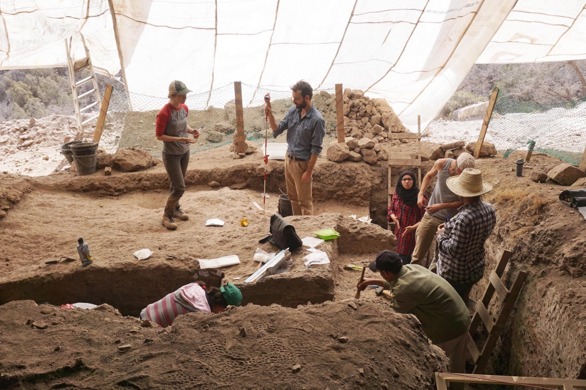 Researchers dig for artifacts in waist-high trenches under a tarp that protects them from the sun