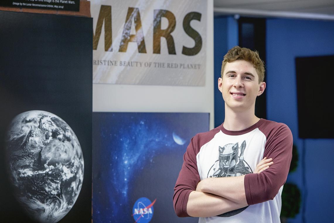young man standing with arms crossed next to a photo of the planet mars UA undergraduate student Gordon Downs is helping develop new ways to analyze data from the NASA Curiosity mars rover as a collaborator on the Mars Science Laboratory science team.