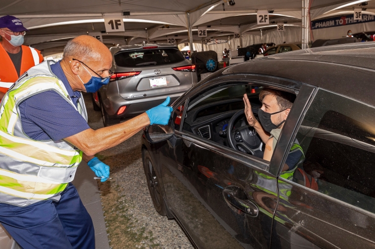 a man wearing a mask gives a thumb up sign to a young man in his car at the university's vaccination site